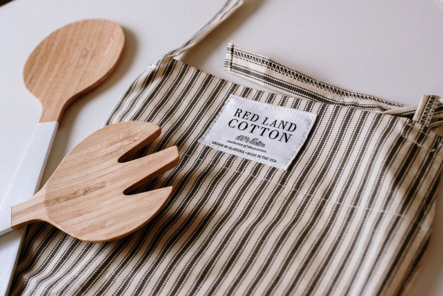 A black and white striped apron with a wooden spoon and a fork with the words Red Ground Cotton on the side