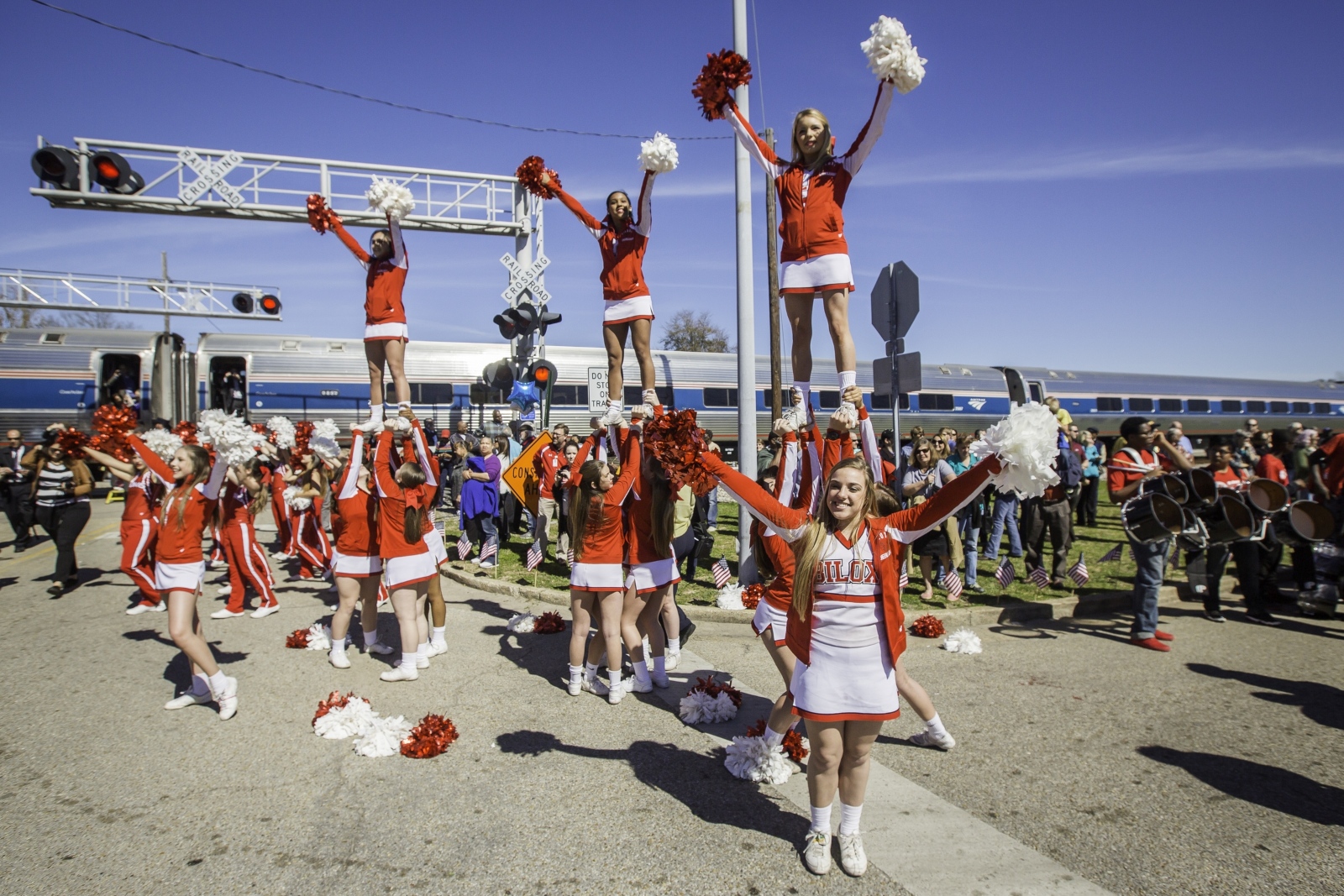 Cheerleaders dressed in red celebrated in front of an Amtrak train during a crowded station event.